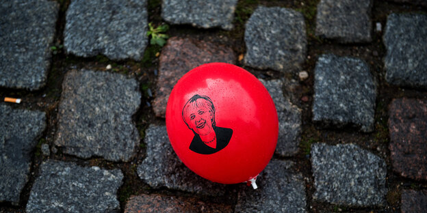 Ein roter Luftballon mit dem Porträt Merkels liegt auf grauem Kopfsteinpflaster Ein roter Luftballon mit dem Porträt Merkels liegt auf grauem Kopfsteinpflaster