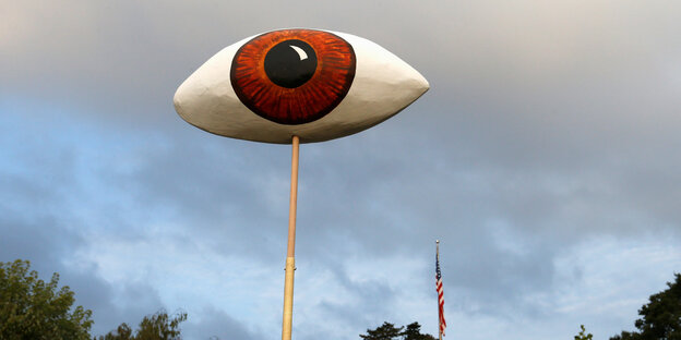 Ein Pappmaché-Auge ragt in den Himmel, weit dahinter eine USA-Fahne Ein Pappmaché-Auge ragt in den Himmel, weit dahinter eine USA-Fahne