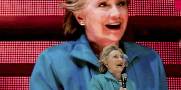 Clinton vor einer Leinwand, die ein Bild von ihr zeigt Clinton vor einer Leinwand, die ein Bild von ihr zeigt