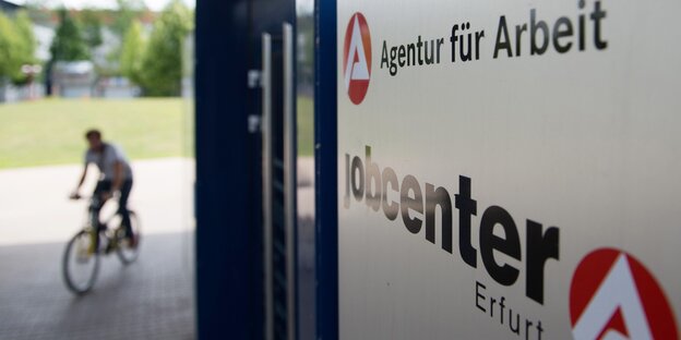 Rechts im Vordergrund des Schild eines Jobcenters, links verschwommen im Hintergrund ein Mensch auf einem Fahrrad Rechts im Vordergrund des Schild eines Jobcenters, links verschwommen im Hintergrund ein Mensch auf einem Fahrrad
