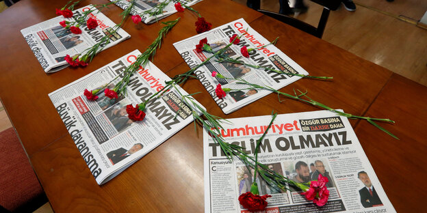 Auf einem Holztisch liegen verschiedene Ausgaben einer Zeitung, auf ihnen mehrere rote Nelken Auf einem Holztisch liegen verschiedene Ausgaben einer Zeitung, auf ihnen mehrere rote Nelken
