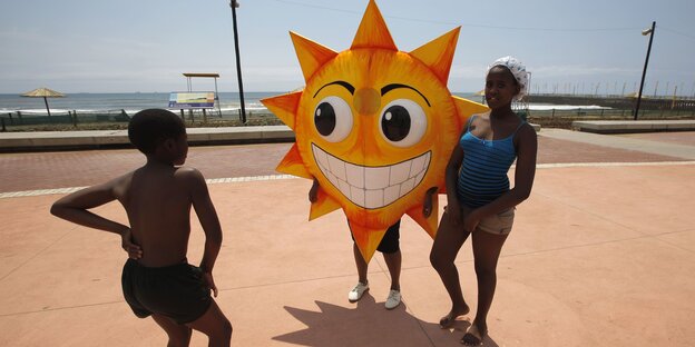 Drei junge Menschen gruppieren sich an einer Strandpromenade um eine Art Sonnenlampion Drei junge Menschen gruppieren sich an einer Strandpromenade um eine Art Sonnenlampion