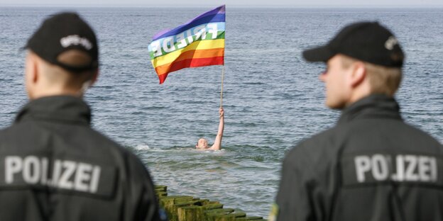 Ein Mann mit einer Regenbogen-Fahne schwimmt im Meer, zwei Polizisten beobachten ihn Ein Mann mit einer Regenbogen-Fahne schwimmt im Meer, zwei Polizisten beobachten ihn