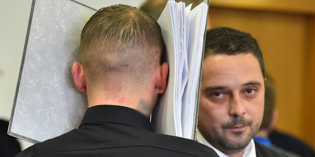 Ein Mann versteckt sein Gesicht hinter einem Aktenordner. Neben ihm steht ein anderer Mann Ein Mann versteckt sein Gesicht hinter einem Aktenordner. Neben ihm steht ein anderer Mann