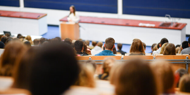 Studierende sitzen in einem Hörsaal, eine weiße Frau spricht Studierende sitzen in einem Hörsaal, eine weiße Frau spricht