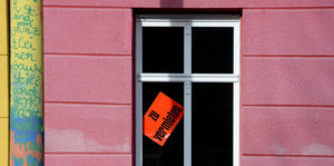 Im Fenster eines Hauses mit rosa gestrichener Fassade steckt ein roter Zettel: Zu vermieten steht darauf