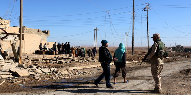 Ein Soldat steht auf einer Straße, zwei Zivilisten laufen an ihm vorbei, im Hintergrund zerstörte Gebäude