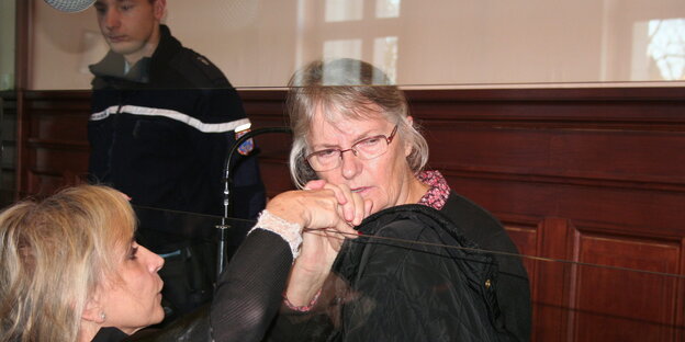 Eine Frau sitzt hinter einer Glasscheibe und blickt zu Boden, eine andere hält ihr tröstend die Hand Eine Frau sitzt hinter einer Glasscheibe und blickt zu Boden, eine andere hält ihr tröstend die Hand