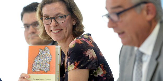 Eine Frau mit Brille sitzt zwischen zwei Männern und hält ein Buch in die Kamera Eine Frau mit Brille sitzt zwischen zwei Männern und hält ein Buch in die Kamera