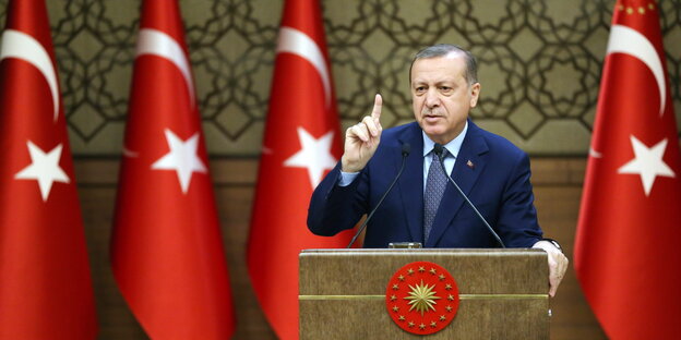 Ein Mann in Anzug steht hinter einem Rednerpult und streckt den Zeigefinger in die Luft. Rechts und links von ihm türkische Flaggen Ein Mann in Anzug steht hinter einem Rednerpult und streckt den Zeigefinger in die Luft. Rechts und links von ihm türkische Flaggen
