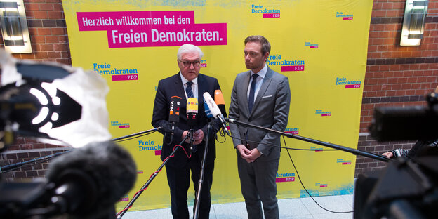 Zwei Männer stehen vor einer gelben Wand. Mikrofone und Kameras sind auf sie gerichtet Zwei Männer stehen vor einer gelben Wand. Mikrofone und Kameras sind auf sie gerichtet