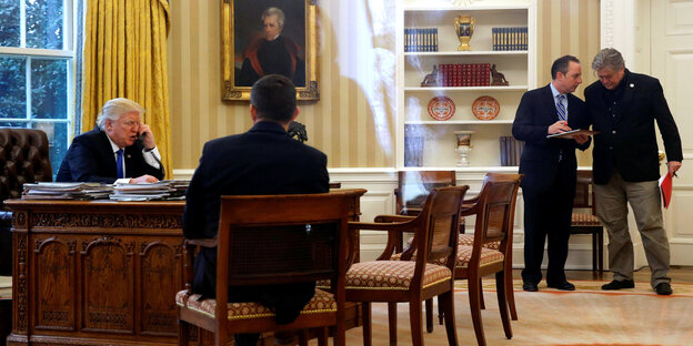 Donald Trump an seinem Schreibtisch im Oval Office. Er telefoniert, während Mitarbeiter sich beraten Donald Trump an seinem Schreibtisch im Oval Office. Er telefoniert, während Mitarbeiter sich beraten