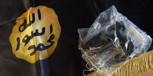 Eine Waffe, eingepackt in Plastik, liegt auf einer IS-Flagge mit arabischen Schriftzeichen Eine Waffe, eingepackt in Plastik, liegt auf einer IS-Flagge mit arabischen Schriftzeichen