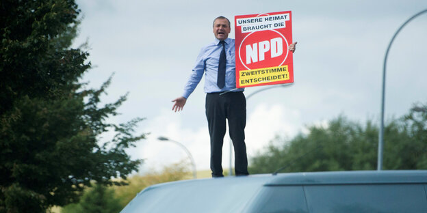 Ein Mann steht auf einem Autodach und hält ein NPD-Plakat im Arm Ein Mann steht auf einem Autodach und hält ein NPD-Plakat im Arm