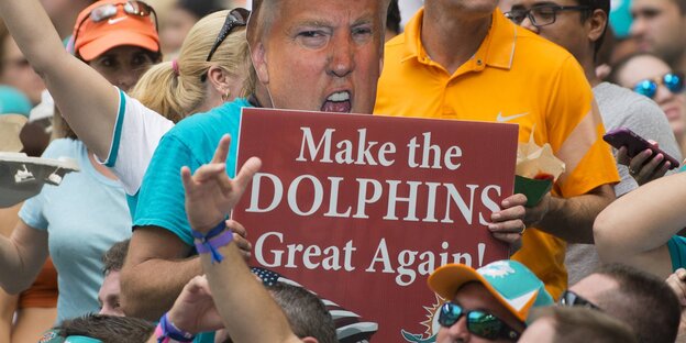 Ein Mann in zwischen Fans hält ein Schild hoch, auf dem Trump zu sehen ist. Darunter der Satz: „Make Dolphins great again" Ein Mann in zwischen Fans hält ein Schild hoch, auf dem Trump zu sehen ist. Darunter der Satz: „Make Dolphins great again"