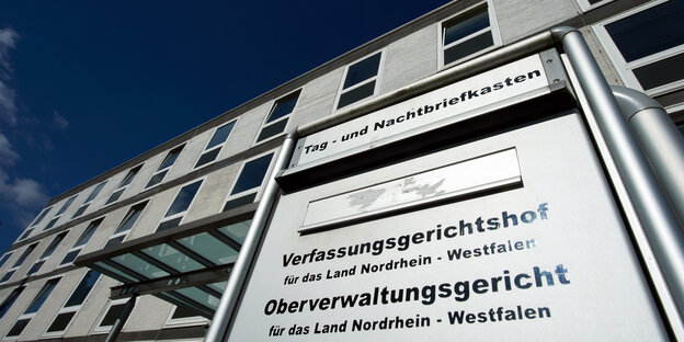 Ein silbernes Schild mit schwarzer Aufschrift vor einem grauen Gebäude Ein silbernes Schild mit schwarzer Aufschrift vor einem grauen Gebäude