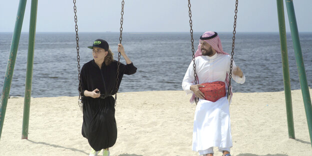 Eine Frau im langen schwarzen Kleid und Käppi und ein Mann in traditionell saudischer Kleidung sitzen am Strand nebeneinander auf Schaukeln Eine Frau im langen schwarzen Kleid und Käppi und ein Mann in traditionell saudischer Kleidung sitzen am Strand nebeneinander auf Schaukeln