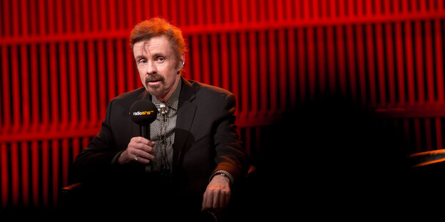 T.C. Boyle sitzt vor rotem Hintergrund und spricht in ein Mikrofon T.C. Boyle sitzt vor rotem Hintergrund und spricht in ein Mikrofon