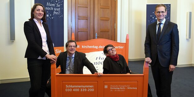 Bürgermeister Müller mit Kirchentagspräsidentin Aus der Au, Landesbischof Dröge und Kirchentags-Generalsekretärin Ueberschär osieren für ein Foto vor einem Bett Bürgermeister Müller mit Kirchentagspräsidentin Aus der Au, Landesbischof Dröge und Kirchentags-Generalsekretärin Ueberschär osieren für ein Foto vor einem Bett