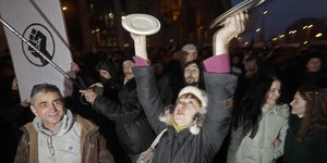 Eine Frau schlägt in einer Menge von Demonstranten zwei Topfdeckel gegeneinander