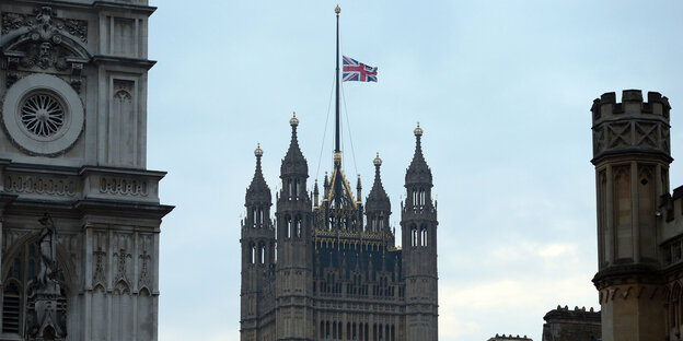 Die Spitze eines pompösen, rechteckigen Gebäudes mit der britischen Flagge darauf, die auf Halbmast weht Die Spitze eines pompösen, rechteckigen Gebäudes mit der britischen Flagge darauf, die auf Halbmast weht
