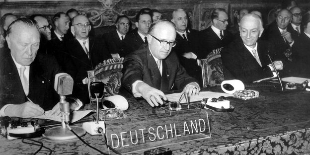 Ein schwarz-weiß-Bild: Drei ältere Männer sitzen nebeneinander und unterzeichnen Dokumente Ein schwarz-weiß-Bild: Drei ältere Männer sitzen nebeneinander und unterzeichnen Dokumente