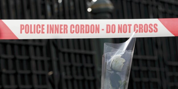 Ein rot-weißes Absperrband, auf dem „Police inner cordon - do not cross“ steht, davor eine weiße Rose in einer durchsichtigen Plastikhülle Ein rot-weißes Absperrband, auf dem „Police inner cordon - do not cross“ steht, davor eine weiße Rose in einer durchsichtigen Plastikhülle