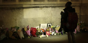 Blumensträuße lehnen an einer Mauer, davor die schwarze Silouette eines Menschen