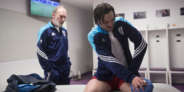 Ein Fußballspieler hält sich in einer Kabine einen Eisbeutel ans Knie, hinter ihm steht ein älterer Herr im Trainingsanzug Ein Fußballspieler hält sich in einer Kabine einen Eisbeutel ans Knie, hinter ihm steht ein älterer Herr im Trainingsanzug