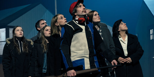 Eine Gruppe von Menschen steht vor einem Bühnenbild mit blauen Häusern Eine Gruppe von Menschen steht vor einem Bühnenbild mit blauen Häusern