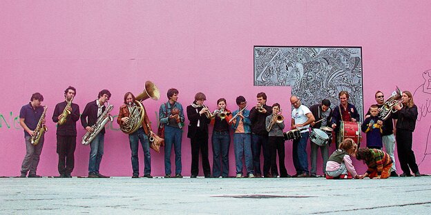 16 Musiker mit Blasinstrumenten und Schlagzeugen vor einer rosa Wand. 16 Musiker mit Blasinstrumenten und Schlagzeugen vor einer rosa Wand.