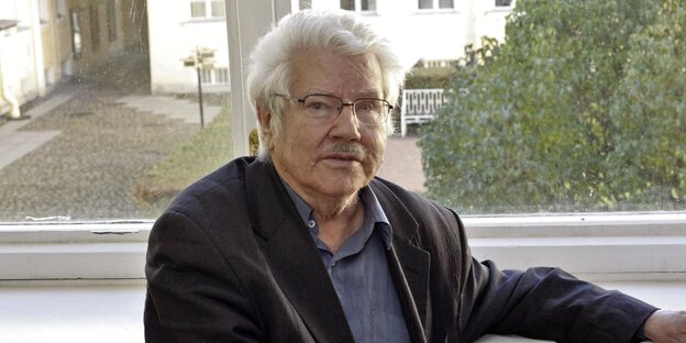 Ein Foto von einem älteren Mann mit grau-weißen Haaren und Brille, der vor einem Fenster sitzt Ein Foto von einem älteren Mann mit grau-weißen Haaren und Brille, der vor einem Fenster sitzt