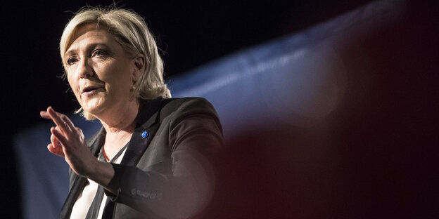 Marine Le Pen steht in Redner-Pose vor nachtschwarzem Hintergrund, nur ein Lichtstrahl fällt hinter ihrem Nacken durchs Bild Marine Le Pen steht in Redner-Pose vor nachtschwarzem Hintergrund, nur ein Lichtstrahl fällt hinter ihrem Nacken durchs Bild