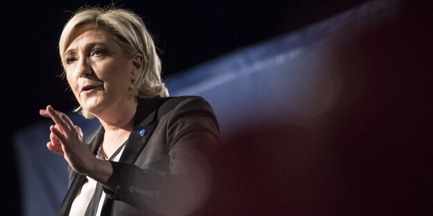 Marine Le Pen steht in Redner-Pose vor nachtschwarzem Hintergrund, nur ein Lichtstrahl fällt hinter ihrem Nacken durchs Bild