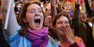 Zwei Frauen, eine im hellblauen, eine im rosa Macron-T-Shirt jubeln, strahlen und eine schwenkt ein Fähnchen
