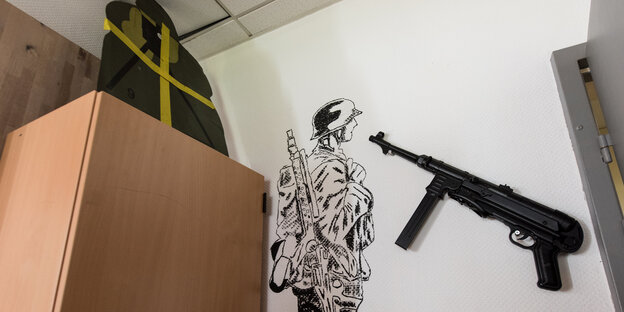 Links ein Schrank, daneben ein an die Wand gezeichneter Soldat und eine Maschinenpistole. Links ein Schrank, daneben ein an die Wand gezeichneter Soldat und eine Maschinenpistole.