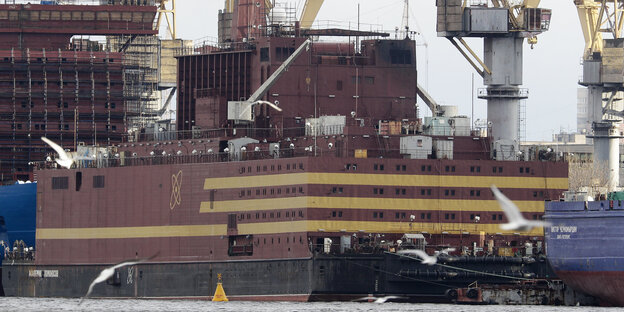 Ein großes Schiff in rotbraun mit beigen Streifen, dahinter Kräne