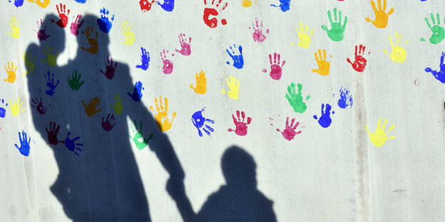 Auf einer Wand, die mit bunten Handabdrücken bemalt ist, sieht man die Schatten von einer erwachsenen Person mit zwei Kindern Auf einer Wand, die mit bunten Handabdrücken bemalt ist, sieht man die Schatten von einer erwachsenen Person mit zwei Kindern