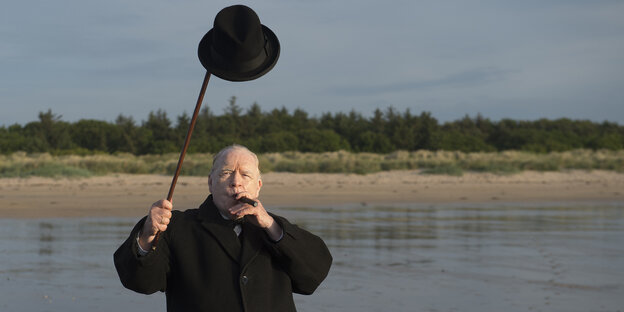 Brian Cox als Winston Churchill am Strand, eine Zigarre im Mund und seinen Bowler mit seinem Gehstock über dem Kopf haltend Brian Cox als Winston Churchill am Strand, eine Zigarre im Mund und seinen Bowler mit seinem Gehstock über dem Kopf haltend