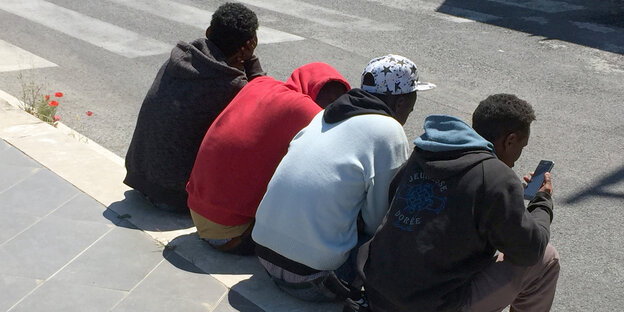 Vier junge Flüchtlinge sitzen nebeneinander auf einer Bordsteinkante, die Rücken gekrümmt. Einer spielt mit seinem Handy Vier junge Flüchtlinge sitzen nebeneinander auf einer Bordsteinkante, die Rücken gekrümmt. Einer spielt mit seinem Handy