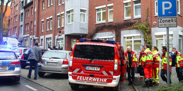 Ein Feuerwehrauto und mehrere Feuerwehrleute und andere Menschen stehen vor Reihenhäusern Ein Feuerwehrauto und mehrere Feuerwehrleute und andere Menschen stehen vor Reihenhäusern