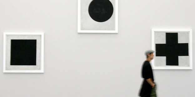 Eine Person läuft an einer Wand in einer Galerie vorbei, an der drei Bilder von Malewitsch hängen: ein schwarzes Quadrat, ein schwarzer Kreis und ein schwarzes Kreuz. Eine Person läuft an einer Wand in einer Galerie vorbei, an der drei Bilder von Malewitsch hängen: ein schwarzes Quadrat, ein schwarzer Kreis und ein schwarzes Kreuz.