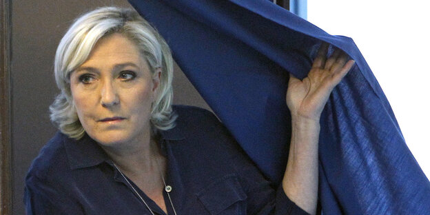Marine Le Pen steht neben einem blauen Vorhang und zieht ihn halb über sich Marine Le Pen steht neben einem blauen Vorhang und zieht ihn halb über sich