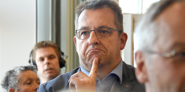 Ein Mann hält seinen Zeigefinger an sein Kinn und guckt fragend, umringt von anderen Menschen Ein Mann hält seinen Zeigefinger an sein Kinn und guckt fragend, umringt von anderen Menschen