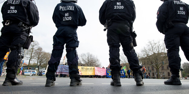 Polizisten in Montur stehen auf einer Straße. Am Horizont sind Demo-Banner zu sehen. Polizisten in Montur stehen auf einer Straße. Am Horizont sind Demo-Banner zu sehen.