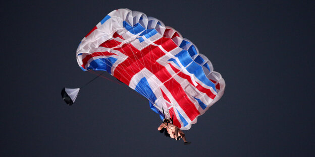 Ein als die Queen verkleideter Schauspieler hängt an einem Fallschirm mit britischem Flaggenmotiv in der Luft Ein als die Queen verkleideter Schauspieler hängt an einem Fallschirm mit britischem Flaggenmotiv in der Luft