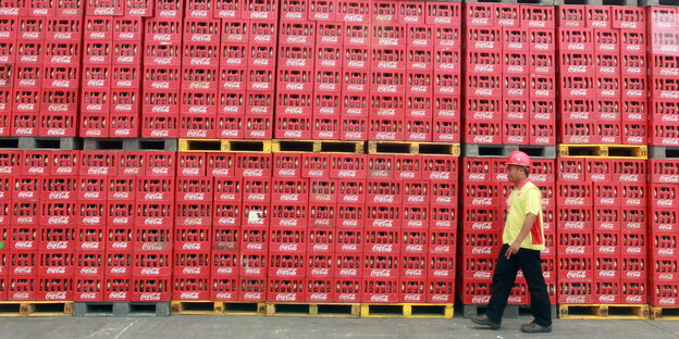 Ein Mann mit Helm geht an einer Wand aus gestapelten Coca-Cola-Kästen vorbei Ein Mann mit Helm geht an einer Wand aus gestapelten Coca-Cola-Kästen vorbei