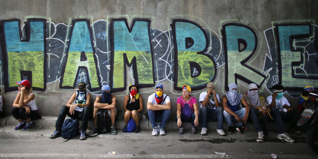 Menschen sitzen vor einer Wand mit der Aufschrift "HAMBRE" Menschen sitzen vor einer Wand mit der Aufschrift "HAMBRE"