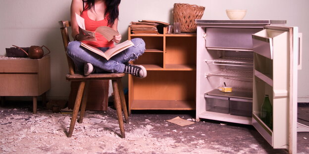 Eine Frau sitzt mit Büchern und Heften auf einem Stuhl neben einem leeren, geöffneten Kühlschrank Eine Frau sitzt mit Büchern und Heften auf einem Stuhl neben einem leeren, geöffneten Kühlschrank