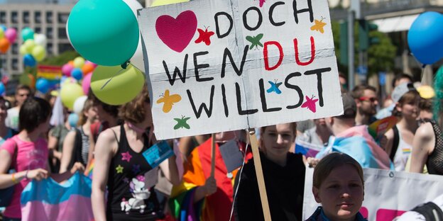 Eine Frau neben einen Schild, auf dem „<3 doch wen du willst" steht, dahinter eine Gruppe Menschen Eine Frau neben einen Schild, auf dem „<3 doch wen du willst" steht, dahinter eine Gruppe Menschen